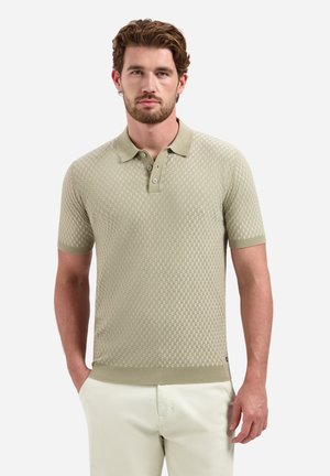 Man met baard die een beige gestructureerd poloshirt met korte mouwen en lichtgekleurde broek draagt, staat met handen deels in de zakken.