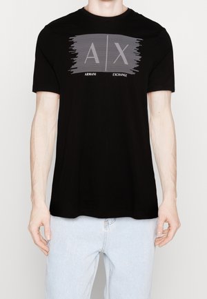 Homme portant un T-shirt noir Armani Exchange avec un grand logo A|X en rayures grises, associé à un jean bleu clair, debout devant un fond uni.