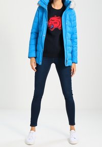 Doudoune bleu vif avec capuche en fausse fourrure, sweat-shirt noir avec un motif rouge, jean skinny foncé et baskets blanches.