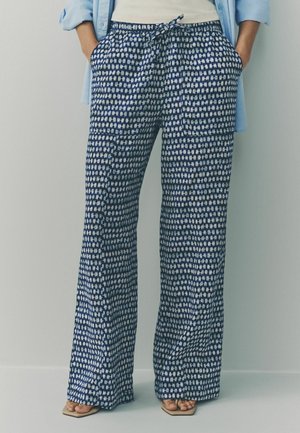 Persona con pantalones anchos de piernas anchas con estampado azul y bolsillos, camisa azul claro y sandalias beige con punta cuadrada, de pie contra una pared lisa.