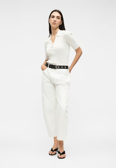 Γυναίκα με μακριά σκουρόχρωμα μαλλιά, φοράει λευκό κοντομάνικο τοπ, λευκά παντελόνια με μαύρη ζώνη και μαύρα σανδάλια με τακούνι, στέκεται με τα χέρια στις τσέπες.