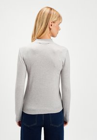 Kobieta z blond włosami ubrana w jasnoszary sweter z długim rękawem i ciemnoniebieskie dżinsy, odwrócona plecami na tle jednolitego białego tła.