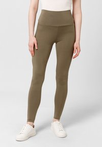 Oliwkowe legginsy z wysokim stanem, wykonane z elastycznego materiału, charakteryzujące się gładką powierzchnią i dopasowanym krojem, w połączeniu z białymi sneakersami.