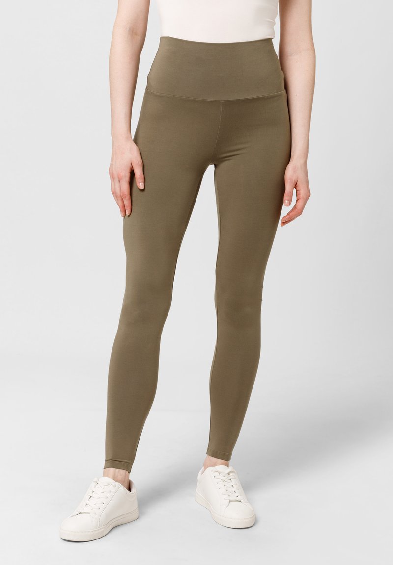 Oliwkowe legginsy z wysokim stanem, wykonane z elastycznego materiału, charakteryzujące się gładką powierzchnią i dopasowanym krojem, w połączeniu z białymi sneakersami.