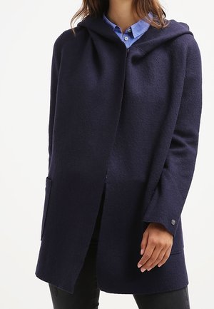 Vrouw draagt een donkerblauwe wollen jas met capuchon over een lichtblauw overhemd met knopen, staand tegen een effen lichte achtergrond.