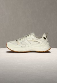 Λευκό αθλητικό παπούτσι Bally με χρυσές λεπτομέρειες, παχιά σόλα, πάνελ από δίχτυ και δέρμα, και επάνω λαβή στην πίσω πλευρά, τοποθετημένο σε επίπεδη επιφάνεια με μπεζ φόντο.