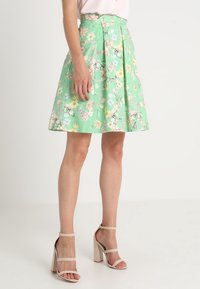 Jupe verte à fleurs avec des plis, présentant un motif vibrant de fleurs roses, bleues et jaunes. Associée à des sandales à talons hauts nudes.