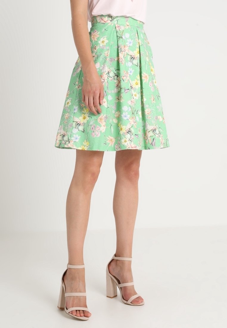 Jupe verte à fleurs avec des plis, présentant un motif vibrant de fleurs roses, bleues et jaunes. Associée à des sandales à talons hauts nudes.