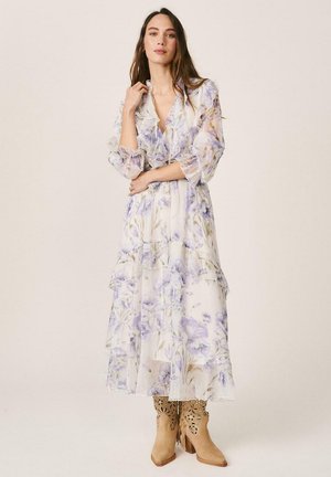 Mujer con un largo vestido floral transparente con detalles de volantes y botas de vaquero de ante color marrón claro con recortes, posando frente a un fondo liso.