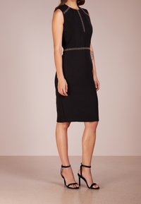 Nainen pukeutuneena hihattomaan mustaan polvipitkään mekkoon, jossa on kultaisia yksityiskohtia, yhdistettynä mustiin korkokenkiin neutraalia taustaa vasten.