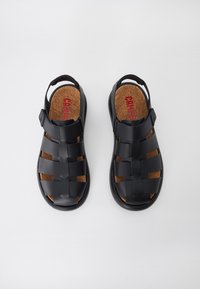 Black sandals featuring woven synthetic straps, a cork insole, and an adjustable backstrap for secure fit. Flat sole design.