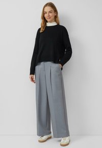 Schwarzer Strickpullover mit geripptem Saum, kombiniert mit hellgrauen, plissierten Hosen mit weitem Bein und Nadelstreifen. Schuhe: weiße Sneaker mit beigen Akzenten.