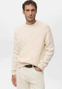 Jasnobeżowy sweter z dzianiny z wzorem warkocza, okrągłym dekoltem i ściągaczami na mankietach, zestawiony z jasno kolorowymi spodniami i czarnym paskiem.