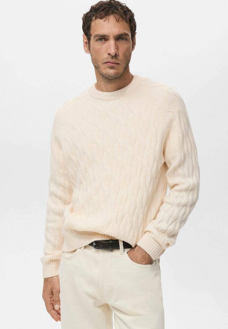 Jasnobeżowy sweter z dzianiny z wzorem warkocza, okrągłym dekoltem i ściągaczami na mankietach, zestawiony z jasno kolorowymi spodniami i czarnym paskiem.