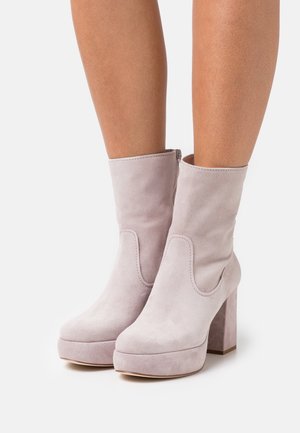 Lyserøde ankelstøvler i ruskind med plateau og blokhæl båret på bare ben mod hvid baggrund.