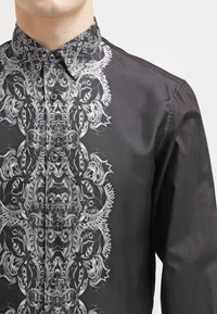 Camicia nera a maniche lunghe con un intricato motivo bianco sulla parte anteriore, con colletto classico e chiusura a bottoni.