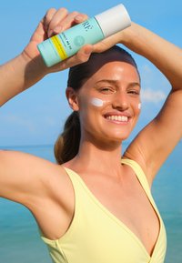 Bottiglia di crema solare con un'etichetta turchese e cappuccio bianco tenuta da una persona in un costume da bagno giallo, con crema bianca sul viso. Sfondo luminoso.