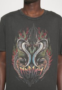 Camiseta de algodón negra con un estampado gráfico de dos serpientes y alas, con llamas rojas y verdes y acentos plateados. Diseño clásico de cuello redondo.