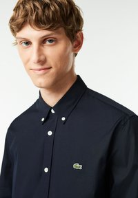 Camicia button-up in cotone blu navy con colletto a punta, dotata di bottoni bianchi e un piccolo logo di coccodrillo verde sul lato sinistro del petto.