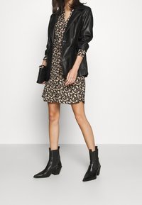 Blazer de piel negra sobre un vestido de estampado floral. Botines negros con punta afilada y tacón grueso. Sosteniendo un pequeño bolso negro tipo clutch.