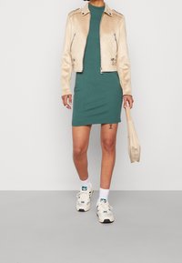 Veste de motard en cuir beige sur une robe verte moulante, associée à des baskets blanches et sarcelle. Tenant un petit sac beige.