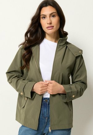 Kvinde med langt mørkt hår iført en åben olivengrøn jakke over en hvid skjorte og blå jeans, stående foran en ensfarvet baggrund.