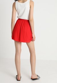 Roter, ausgestellt Minirock mit einem elastischen Bund, kombiniert mit einem weißen Tanktop. Das Model trägt schwarz-weiße Slide-Sandalen.