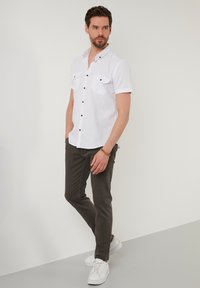Camisa blanca de manga corta con bolsillos abotonados, botones marrones y pantalones grises. Modelo que lleva zapatillas blancas, de pie sobre una superficie clara.