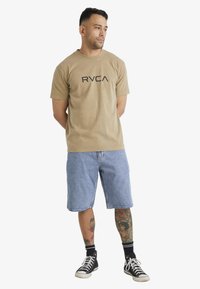 Khaki puuvillainen T-paita, jossa on musta "RVCA" -logo, yhdistettynä vaaleisiin farkkushortseihin ja mustiin korkeakärkisiin lenkkareihin. Henkilö seisoo kädet rentoina.
