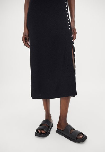 Schwarzes gestricktes Midi-Kleid mit seitlichen Schlitzen und weißen Knopfakzenten; kombiniert mit klobigen schwarzen Rutschsandalen mit einer dicken Sohle.