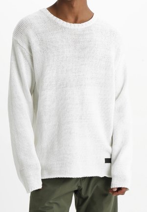 White knitted sweater with a round neckline, long sleeves, loose fit, and a black label detail at the hem. Textured and lightweight design.
