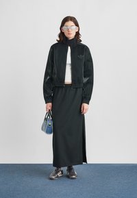 Casaco atlético preto com fecho éclair e logótipo no peito, combinado com uma saia preta justa com uma racha lateral. Acessórios incluem uma mala azul e óculos de sol.