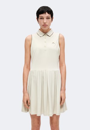 Mujer joven con cabello corto y negro que lleva un vestido beige sin mangas con cuello, botones y falda plisada, de pie contra un fondo liso.