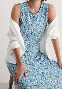 Kobieta siedząca na drewnianym stołku, ubrana w niebieską, kwiecistą sukienkę bez rękawów, z białą koszulą zarzuconą na ramionach.