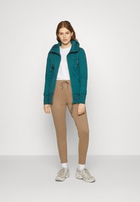 Teal zip-up hettegenser med snor, sammen med beige joggebukser. Fottøyet inkluderer lysegrå sneakers. Modellen står mot en hvit bakgrunn.