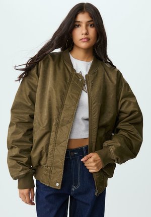 Young woman with long dark hair wearing olive-green bomber jacket, cropped light grey shirt with star detail, and high-waisted dark blue jeans.