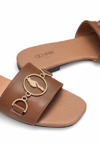 Brązowe klapki na płaskiej podeszwie, ozdobione dużym złotym okrągłym emblematem oraz małym złotym akcentem na pasku.