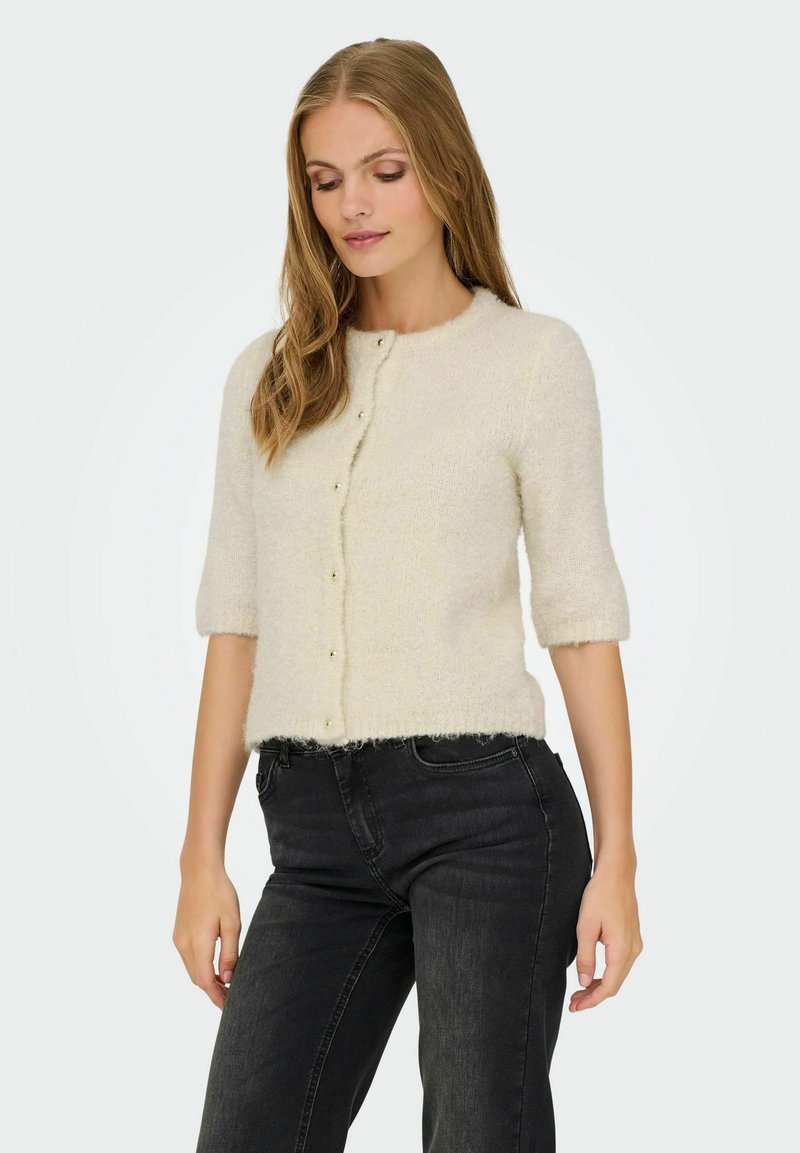 Femme aux longs cheveux blonds portant un cardigan crémeux à boutons et un jean noir taille haute, regardant vers le bas avec les mains détendues.