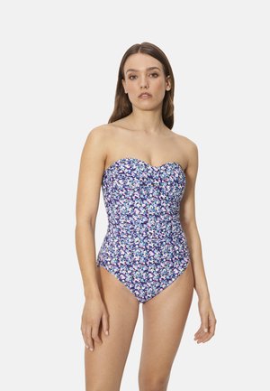 Mujer de pie con un traje de baño de una pieza sin tirantes con un patrón floral azul, blanco y púrpura sobre un fondo liso.