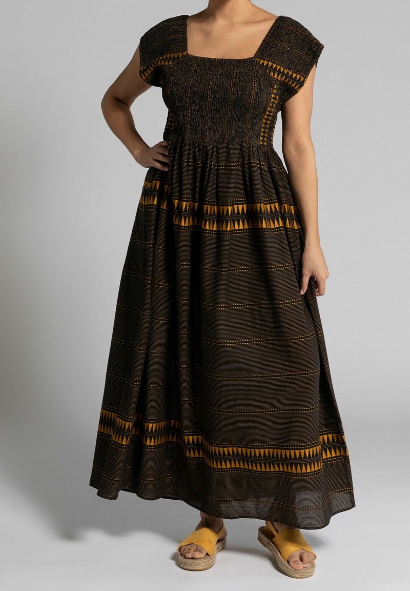 Femme portant une robe maxi noire et orange à motifs, manches courtes, et des espadrilles jaunes, debout avec une main sur la hanche.