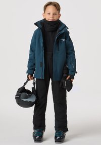 Strój narciarski: turkusowa wodoodporna kurtka, czarna termiczna bluza, czarne spodnie narciarskie, czarne rękawice, buty narciarskie, kask trzymany w ręce. Zapięcia na zamek błyskawiczny i logo.