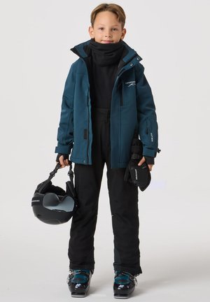 Skibeklædning: turkis vandtæt jakke, sort termooverdel, sorte skibukser, sorte handsker, skistøvler, hjelm i hånden. Har lynlåse og logo.