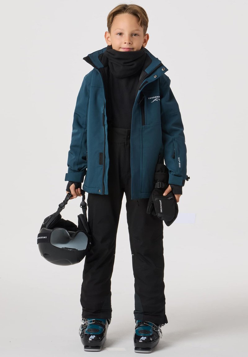 Strój narciarski: turkusowa wodoodporna kurtka, czarna termiczna bluza, czarne spodnie narciarskie, czarne rękawice, buty narciarskie, kask trzymany w ręce. Zapięcia na zamek błyskawiczny i logo.