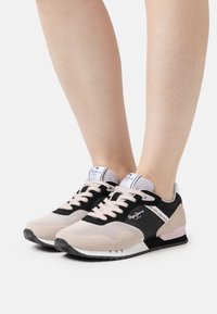 Sneakers i beige, svart och ljusrosa. Materialet inkluderar mesh och mocka. Har vita detaljer, svarta snören och branding på sidan.