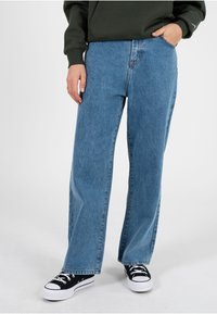 Persoană purtând blugi albaștri cu picioare largi, adidași alb-negru și un pulover verde închis, stând cu o mână în buzunar.