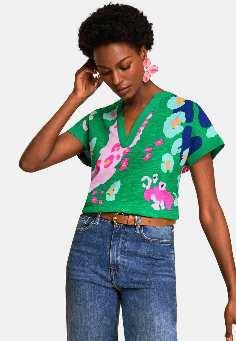 Lühikeste lokkidega naine kohandab kõrvaklappi, kandes rohelist lillelist ülaosa V-kaelusega ja siniseid teksapükse pruuni vööga valge tausta ees.