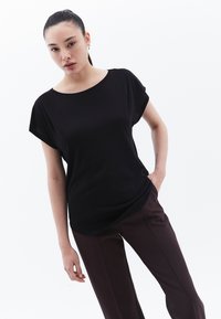 Musta lyhythihainen toppi, jossa on leveä pääntie ja löysä istuvuus, yhdistettynä tummanruskeisiin korkea vyötärön housuihin, taustalla valkoinen tausta.