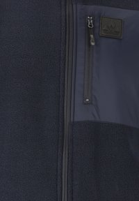 Marineblå fleecejakke med en glat nylonpanel, lynlås brystlomme og struktureret stof. Har et logo-patch på brystet.