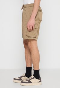 Kaki cargoshorts met een elastische tailleband en trekkoord, voorzien van een enkele zijzak en een relaxte pasvorm. Gemaakt in combinatie met beige en grijze sneakers.