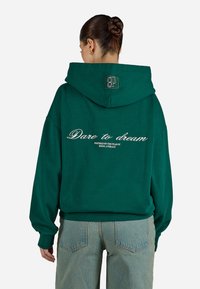 Πράσινη oversized hoodie από μαλακό ύφασμα, με μεγάλη κουκούλα, ριμπ μανσέτες και κείμενο "Dare to dream" εκτυπωμένο στην πλάτη.